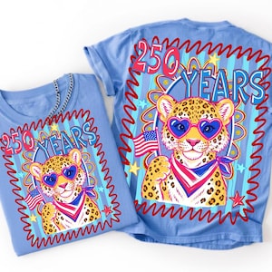 Op de afbeelding: Twee lichtblauwe t-shirts met een luipaard met een hartvormige zonnebril en een Amerikaanse vlag. De shirts hebben de tekst "250 YEARS" in een rood-blauw ontwerp.
