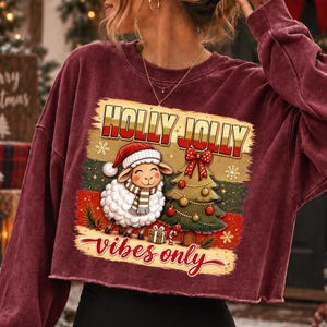 Może przedstawiać: Bordowa bluza z długim rękawem i kr&oacute;tkim krojem z nadrukiem owcy w czapce Mikołaja i szaliku obok udekorowanej choinki. Napis HOLLY JOLLY vibes only wykonany jest czerwoną i ż&oacute;łtą czcionką z akcentami w kształcie płatk&oacute;w śniegu.