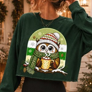 May include: A dark green long-sleeve cropped sweatshirt featuring a graphic of a brown owl wearing a red, white, and green striped knit hat and a green scarf. The owl holds a yellow mug with white marshmallows, set against a snowy background.