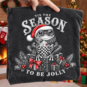 Puede incluir: Una camiseta gris carb&oacute;n con un gr&aacute;fico de una rana con gorro de Pap&aacute; Noel y gafas redondas, sosteniendo una taza de chocolate caliente con nubes. El dise&ntilde;o incluye cajas de regalo apiladas, hojas de acebo y el texto 'TIS THE SEASON TO BE JOLLY'.