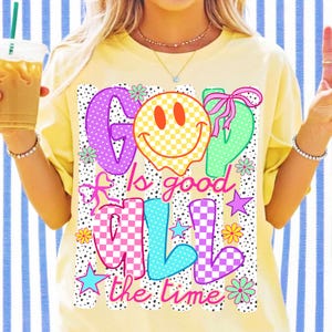 Op de afbeelding: Een lichtgeel t-shirt met een grafische print met de tekst God Is Good All The Time. Het ontwerp bevat een geel geblokt smileygezicht, roze strikken, kleurrijke sterren en bloemen op een achtergrond van zwarte stippen en verticale strepen.