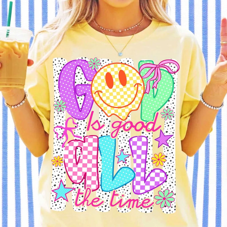 Op de afbeelding: Een lichtgeel t-shirt met een grafische print met de tekst God Is Good All The Time. Het ontwerp bevat een geel geblokt smileygezicht, roze strikken, kleurrijke sterren en bloemen op een achtergrond van zwarte stippen en verticale strepen.