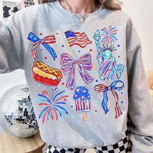 K&ouml;nnte beinhalten: Ein hellgraues Sweatshirt mit patriotischen Illustrationen. Das Design zeigt eine wehende amerikanische Flagge, die Freiheitsstatue, Feuerwerk, Schleifen, einen Hotdog und ein Eis am Stiel, alles in Rot, Wei&szlig; und Blau.