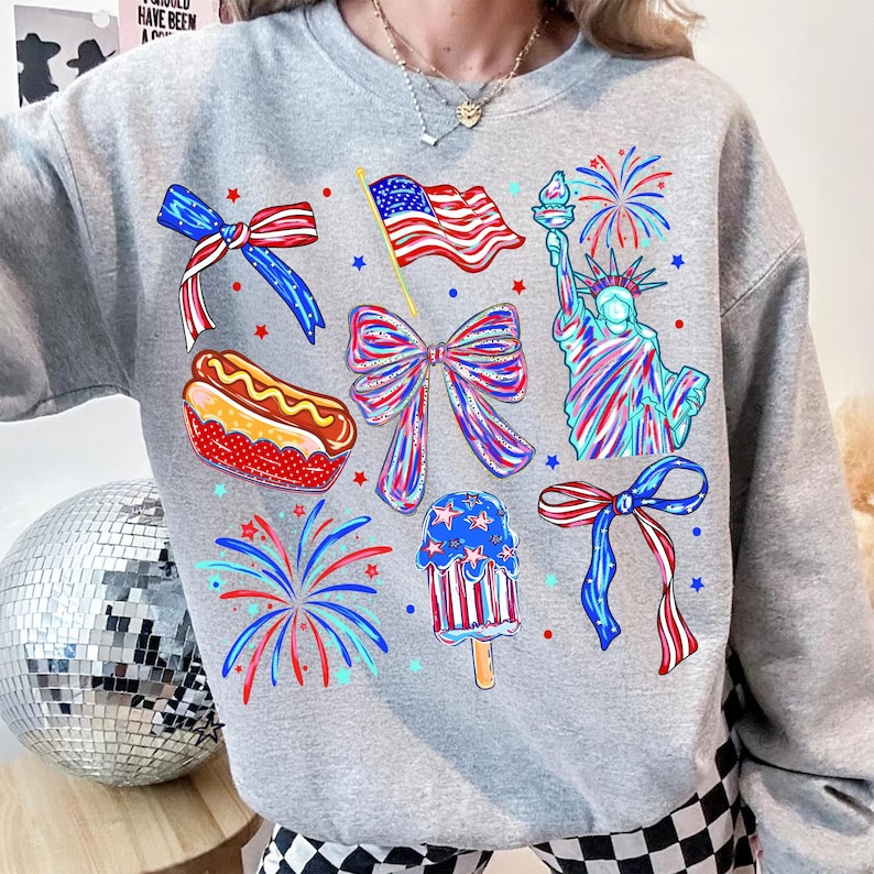 K&ouml;nnte beinhalten: Ein hellgraues Sweatshirt mit patriotischen Illustrationen. Das Design zeigt eine wehende amerikanische Flagge, die Freiheitsstatue, Feuerwerk, Schleifen, einen Hotdog und ein Eis am Stiel, alles in Rot, Wei&szlig; und Blau.