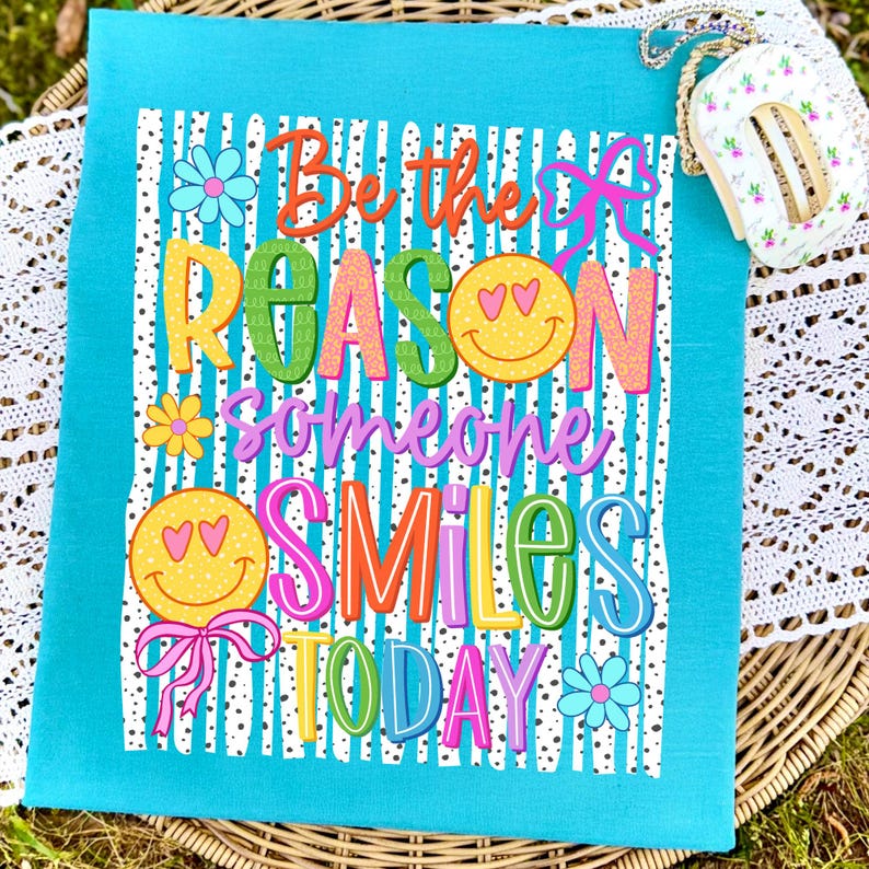 May include: A turquoise kitchen towel with the text "Be the REASON someone SMILES TODAY" in colorful, playful lettering. The design includes smiley faces, flowers, and bows. The towel is likely made of cotton or a similar absorbent material.