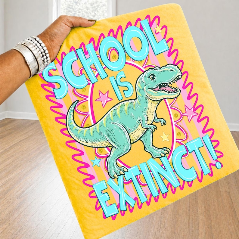 Op de afbeelding: Gele deken met een cartoon dinosaurus en de tekst "SCHOOL IS EXTINCT!" in turquoise. De dinosaurus is teal met gele accenten, tegen een roze en gele achtergrond.