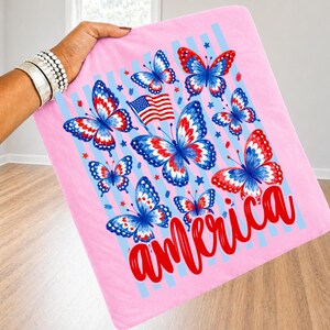 K&ouml;nnte beinhalten: Ein hellrosa Stoffquadrat mit einem Grafikdruck mehrerer Schmetterlinge mit rot-wei&szlig;-blauen Fl&uuml;gelmustern. Eine kleine amerikanische Flagge ist zwischen den Schmetterlingen platziert und das Wort america ist in roter Schreibschrift gedruckt.