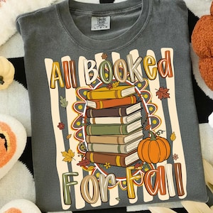 Op de afbeelding: Een antracietgrijs t-shirt met een grafische print van een stapel boeken en een oranje pompoen. De tekst "All Booked For Fall" is gedrukt in een veelkleurig, gestileerd lettertype. Het ontwerp bevat herfstbladeren en een gestreepte achtergrond.