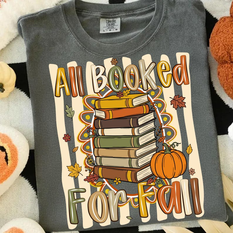 Op de afbeelding: Een antracietgrijs t-shirt met een grafische print van een stapel boeken en een oranje pompoen. De tekst "All Booked For Fall" is gedrukt in een veelkleurig, gestileerd lettertype. Het ontwerp bevat herfstbladeren en een gestreepte achtergrond.