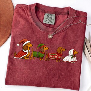 May include: A maroon short-sleeve t-shirt featuring a graphic of four dachshunds dressed in holiday attire, including Santa and elf costumes. The shirt has a Comfort Colors label at the neck and a thin silver chain necklace resting on the fabric.