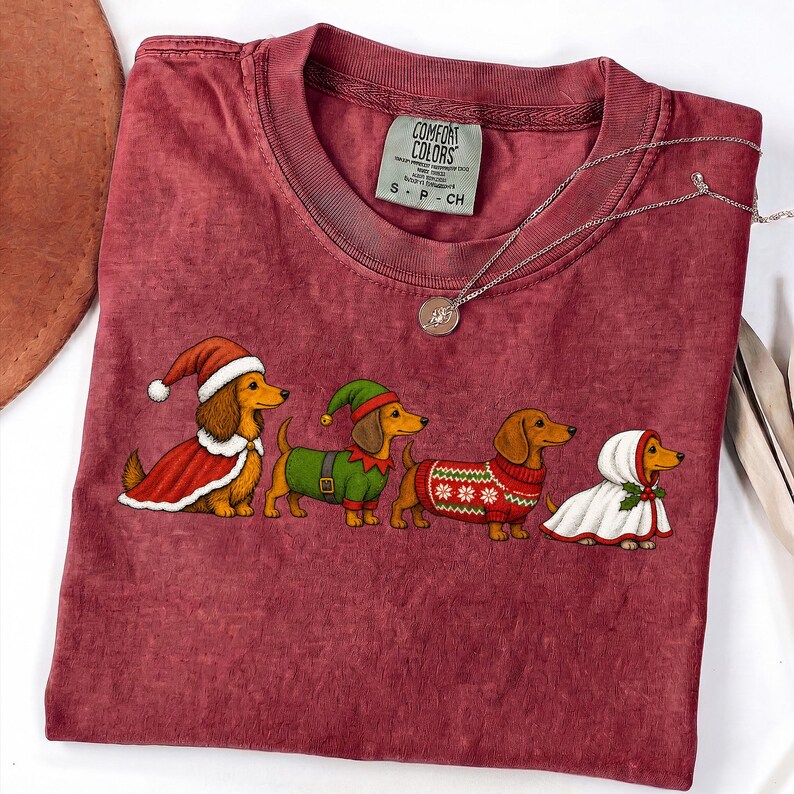 May include: A maroon short-sleeve t-shirt featuring a graphic of four dachshunds dressed in holiday attire, including Santa and elf costumes. The shirt has a Comfort Colors label at the neck and a thin silver chain necklace resting on the fabric.