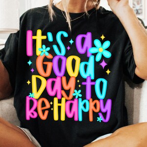 K&ouml;nnte beinhalten: Schwarzes T-Shirt mit einer bunten Grafik, auf der "It's a good day to be happy" steht. Die Grafik zeigt Blumen und Sterne.