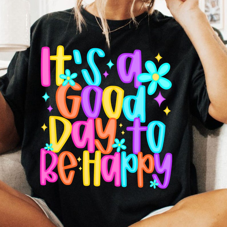 K&ouml;nnte beinhalten: Schwarzes T-Shirt mit einer bunten Grafik, auf der "It's a good day to be happy" steht. Die Grafik zeigt Blumen und Sterne.