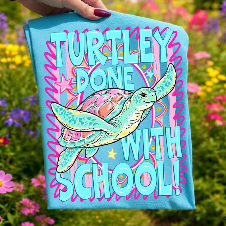 以下が含まれることがあります： 「TURTLEY DONE WITH SCHOOL！」の文字と、漫画のウミガメのイラストが描かれた水色のTシャツ。カメはピンクとオレンジ色の甲羅を持ち、ピンクと青の装飾的な要素に囲まれています。