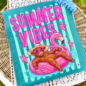 Może przedstawiać: Turkusowa poduszka z r&oacute;żowym napisem "SUMMER VIBES". Ilustracja przedstawia szkocką krowę w okularach przeciwsłonecznych i słomkowym kapeluszu, leżącą na r&oacute;żowym dmuchanym flamingu. R&oacute;żowe gwiazdki i jasnoniebieskie paski dopełniają wz&oacute;r.