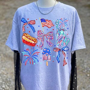 K&ouml;nnte beinhalten: Hellgraues T-Shirt mit patriotischen Illustrationen. Das Design zeigt eine wehende amerikanische Flagge, die Freiheitsstatue, Feuerwerk, Schleifen, einen Hotdog und ein Eis am Stiel, alles in Rot, Wei&szlig; und Blau.