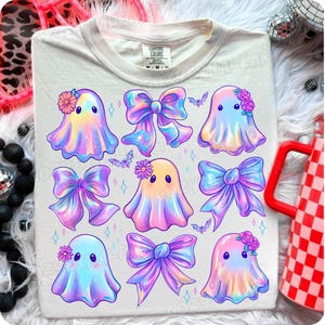 Może przedstawiać: Kremowa koszulka z kolorowym wzorem kreskówkowych duchów i kokard. Duchy są w pastelowych kolorach z kwiatowymi akcentami. Kokardy są w odcieniach fioletu i niebieskiego. Zawiera również małe ilustracje nietoperzy.