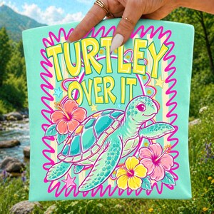Puede incluir: Bolso tote de color verde azulado claro con las palabras "TURTLEY OVER IT" en amarillo y rosa. El bolso presenta una tortuga de dibujos animados con un caparaz&oacute;n turquesa y flores de hibisco rosas y amarillas. El dise&ntilde;o est&aacute; enmarcado por un borde ondulado rosa.
