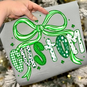 May include: A gray fabric item with a large green bow and the word "CUSTOM" in green and white lettering. The letters have various patterns, including polka dots, shamrocks, and gingham. Small green shamrocks are scattered around the text.