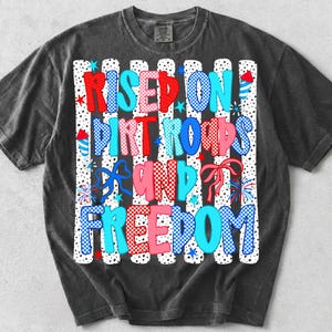 Op de afbeelding: Een antracietgrijs t-shirt met een grafische print met de tekst "RISED ON DIRT ROADS AND FREEDOM" in rood, wit en blauwe ruit- en stippenpatronen, geaccentueerd met illustraties van vuurwerk, sterren en strikken op een gestreepte achtergrond.