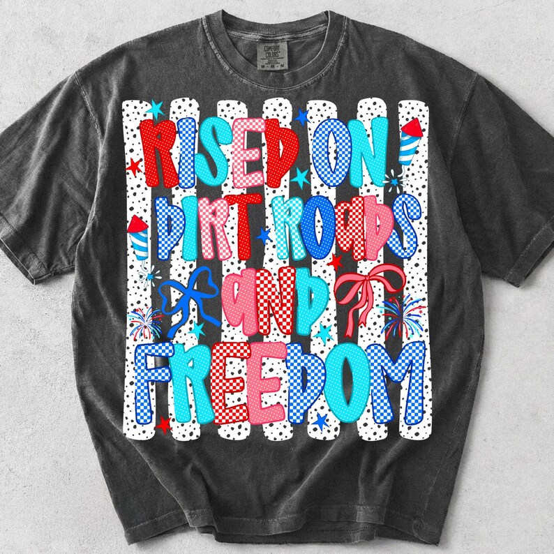 Op de afbeelding: Een antracietgrijs t-shirt met een grafische print met de tekst "RISED ON DIRT ROADS AND FREEDOM" in rood, wit en blauwe ruit- en stippenpatronen, geaccentueerd met illustraties van vuurwerk, sterren en strikken op een gestreepte achtergrond.
