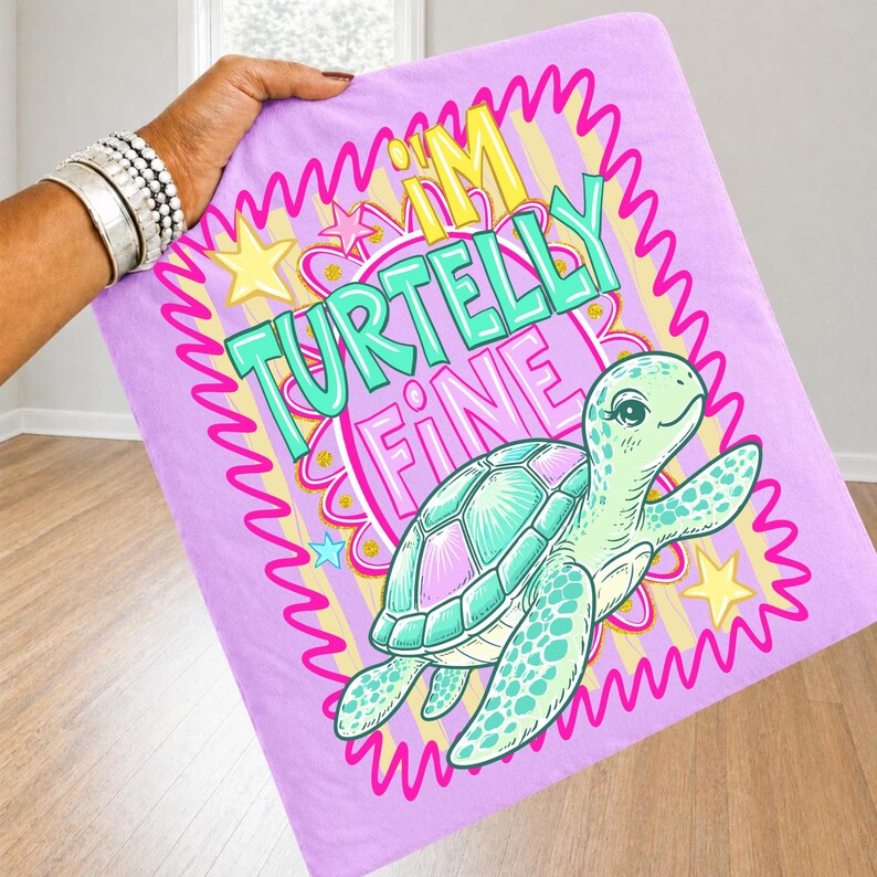 May include: A light purple blanket with a cartoon turtle illustration and the text "I'M TURTELLY FINE". The turtle is green and has a pink and teal shell. The design is framed with a pink wavy border and yellow stars.