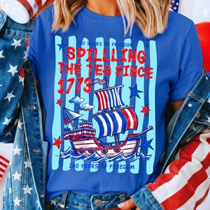 Pu&ograve; includere: Maglietta blu con un design patriottico di una nave rossa, bianca e blu. Il testo recita "Spilling the Tea Since 1773" e "250 Years of Freedom". Il design include stelle e strisce.