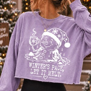 May include: A light purple long-sleeve crop sweatshirt featuring a white graphic of a melting snowman wearing a Santa hat and holding a mug. The text reads, "WINTER'S PACE, LET IT MELT." with a small heart icon below the text on the front.