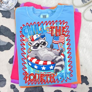 Op de afbeelding: Een lichtblauw t-shirt met een afbeelding van een wasbeer met een zonnebril en een patriotische hoed, liggend in een rood, wit en blauw opblaasbare ring. De tekst luidt "Chill the Fourth Out."