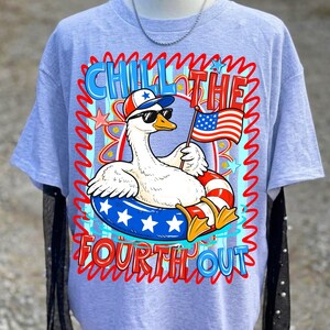 May include: Light blue t-shirt featuring a cartoon goose wearing sunglasses and a patriotic hat, floating in a red, white, and blue inner tube. The shirt has the text "CHILL THE FOURTH OUT" in red and blue.
