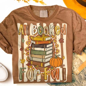 May include: A brown short-sleeve t-shirt featuring a graphic of a stack of six books and an orange pumpkin. The text "All Booked For Fall" is printed in a stylized font above and below the stack, surrounded by autumn leaves and flowers.