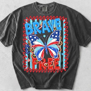 May include: Dark grey t-shirt with a patriotic butterfly design. The butterfly has red, white, and blue wings with stars. The words "BRAVE & FREE" are in blue and red, with a red zigzag border.