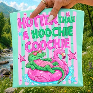 May include: A teal tote bag with pink and green text that reads "HOTTER THAN A HOOCHIE COOCHIE." The bag features a cartoon alligator wearing heart-shaped sunglasses and a flamingo on a pink floatie. Pink and blue stars are scattered around the design.