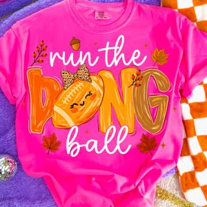 Puede incluir: Una camiseta de color rosa ne&oacute;n brillante con el texto "run the ball" en letra cursiva blanca. El dise&ntilde;o incluye un bal&oacute;n de f&uacute;tbol americano con un lazo de estampado de leopardo, una bellota, hojas de oto&ntilde;o y bayas en tonos naranja, marr&oacute;n y amarillo.