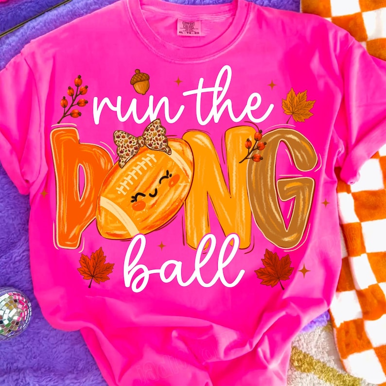 Puede incluir: Una camiseta de color rosa ne&oacute;n brillante con el texto "run the ball" en letra cursiva blanca. El dise&ntilde;o incluye un bal&oacute;n de f&uacute;tbol americano con un lazo de estampado de leopardo, una bellota, hojas de oto&ntilde;o y bayas en tonos naranja, marr&oacute;n y amarillo.
