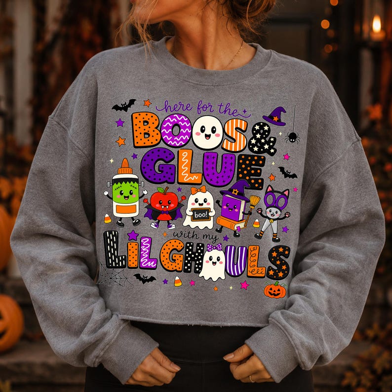 K&ouml;nnte beinhalten: Ein graumeliertes Langarm-Sweatshirt mit einer bunten Halloween-Grafik und dem Text "here for the BOOS & GLUE with my LILGHOULS". Das Design zeigt verschiedene Zeichentrickfiguren wie einen Geist, ein Buch und eine Klebeflasche.