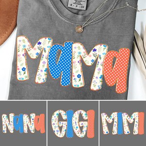 Może przedstawiać: Szara koszulka z napisem "Mama" dużymi, kolorowymi literami. Litery ozdobione są kwiatowymi wzorami, niebieskimi kropkami i pomarańczowymi gwiazdkami. Widoczny jest srebrny naszyjnik z małą zawieszką.
