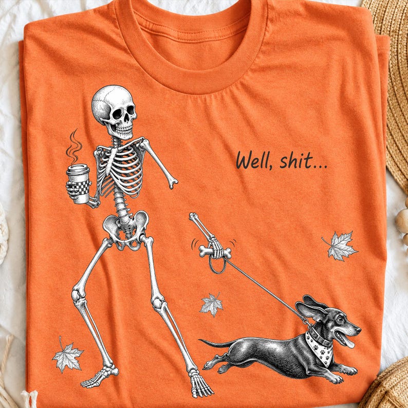 May include: An orange crew neck t-shirt featuring a black and white graphic of a skeleton holding a coffee cup and a bone-shaped leash attached to a dachshund. The text "Well, shit..." is printed above the dog, with scattered maple leaves.