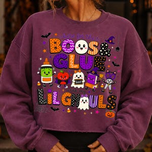 K&ouml;nnte beinhalten: Ein pflaumenfarbenes Sweatshirt mit Rundhalsausschnitt und einem Grafikdruck mit dem Text "here for the BOOS & GLUE with my LIL GHOULS". Das Design zeigt bunte Cartoon-Illustrationen von Kleber, Apfel, Geist, Buch und Katze, umgeben von Sternen.