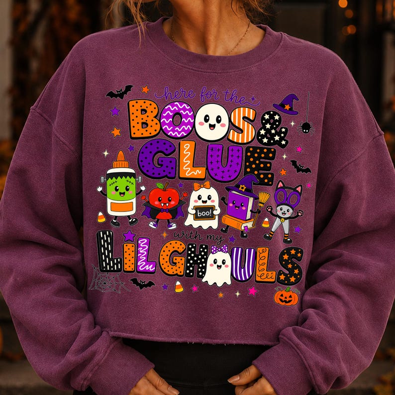 K&ouml;nnte beinhalten: Ein pflaumenfarbenes Sweatshirt mit Rundhalsausschnitt und einem Grafikdruck mit dem Text "here for the BOOS & GLUE with my LIL GHOULS". Das Design zeigt bunte Cartoon-Illustrationen von Kleber, Apfel, Geist, Buch und Katze, umgeben von Sternen.