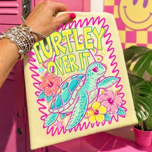 Puede incluir: Una toalla de playa de color amarillo claro con la frase "TURTLEY OVER IT" en amarillo y verde. La toalla presenta una tortuga marina de dibujos animados en turquesa y rosa, rodeada de flores y estrellas rosas y amarillas.
