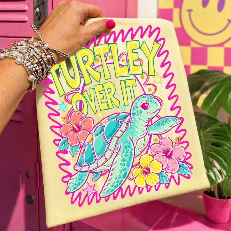 Puede incluir: Una toalla de playa de color amarillo claro con la frase "TURTLEY OVER IT" en amarillo y verde. La toalla presenta una tortuga marina de dibujos animados en turquesa y rosa, rodeada de flores y estrellas rosas y amarillas.