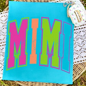 May include: A turquoise tote bag with the word "MIMI" in large, distressed, colorful letters. The letters are pink, orange, and green with a neon outline. A small floral purse with a gold chain is visible.