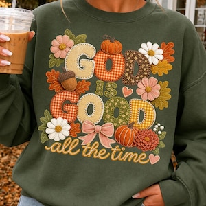 Pu&ograve; includere: Una felpa girocollo verde oliva con un design grafico che riporta il testo "GOD IS GOOD all the time". Le lettere sono riempite con vari motivi, tra cui quadri, fiori e texture in iuta, circondate da zucche, fiori e foglie decorative.