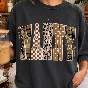 May include: A charcoal grey short-sleeve t-shirt featuring the word NANNY printed in large block letters. Each letter has a unique pattern including camouflage, brown and white checkerboard, leopard print, and white polka dots with small bows.
