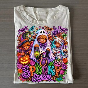 Może przedstawiać: Jasny t-shirt z grafiką przedstawiającą trzy szkockie krowy przebrane na Halloween za czarownicę, ducha i mumię. Wz&oacute;r zawiera dynie, nietoperze, pajęczyny oraz napis "SPOOKY SEASON" wykonany kolorowymi, wzorzystymi literami.