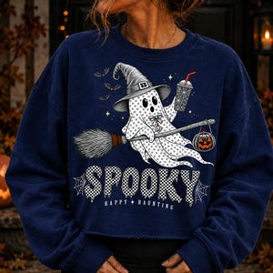 May include: A navy blue sweatshirt featuring a graphic of a ghost wearing a witch hat, holding a drink and a broomstick with a hanging jack-o'-lantern. The text reads SPOOKY HAPPY HAUNTING in a checkered pattern with spider web accents.