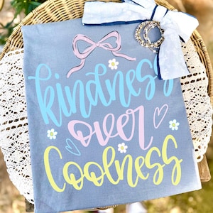 May include: A gray t-shirt with the words "Kindness Over Coolness" in blue, pink, and yellow script. The shirt features a pink bow graphic and small flower and heart accents. A white bow and bracelets are on top of the shirt.