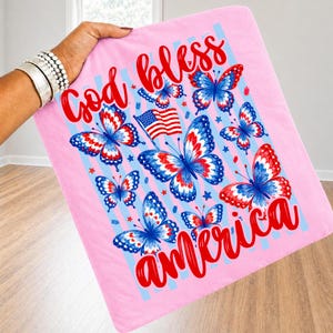 May include: A light pink square fabric item featuring a graphic print of red, white, and blue butterflies, a small American flag, and the red script text God bless America. The design includes light blue vertical stripes and small red stars.
