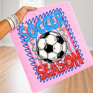 Puede incluir: Manta de f&uacute;tbol rosa con un bal&oacute;n de f&uacute;tbol en blanco y negro. La manta tiene las palabras "SOCCER SEASON" en azul y rojo. La manta est&aacute; sostenida por una persona.